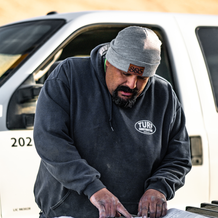 A Turf Construction employee wearing a company branded sweatshirt looks over a set of civil plans on a job site.