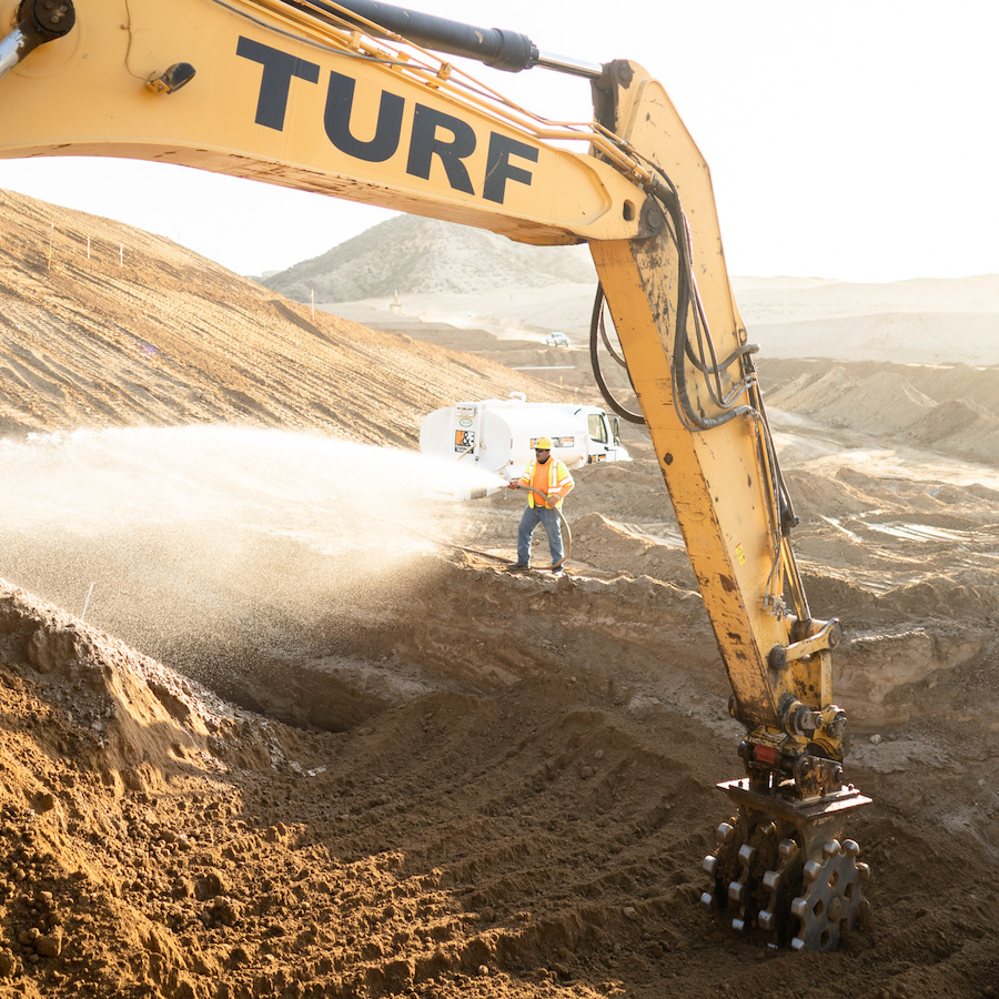 Turf Construction excavator with compactor wheel and Turf Construction employee with water hose.